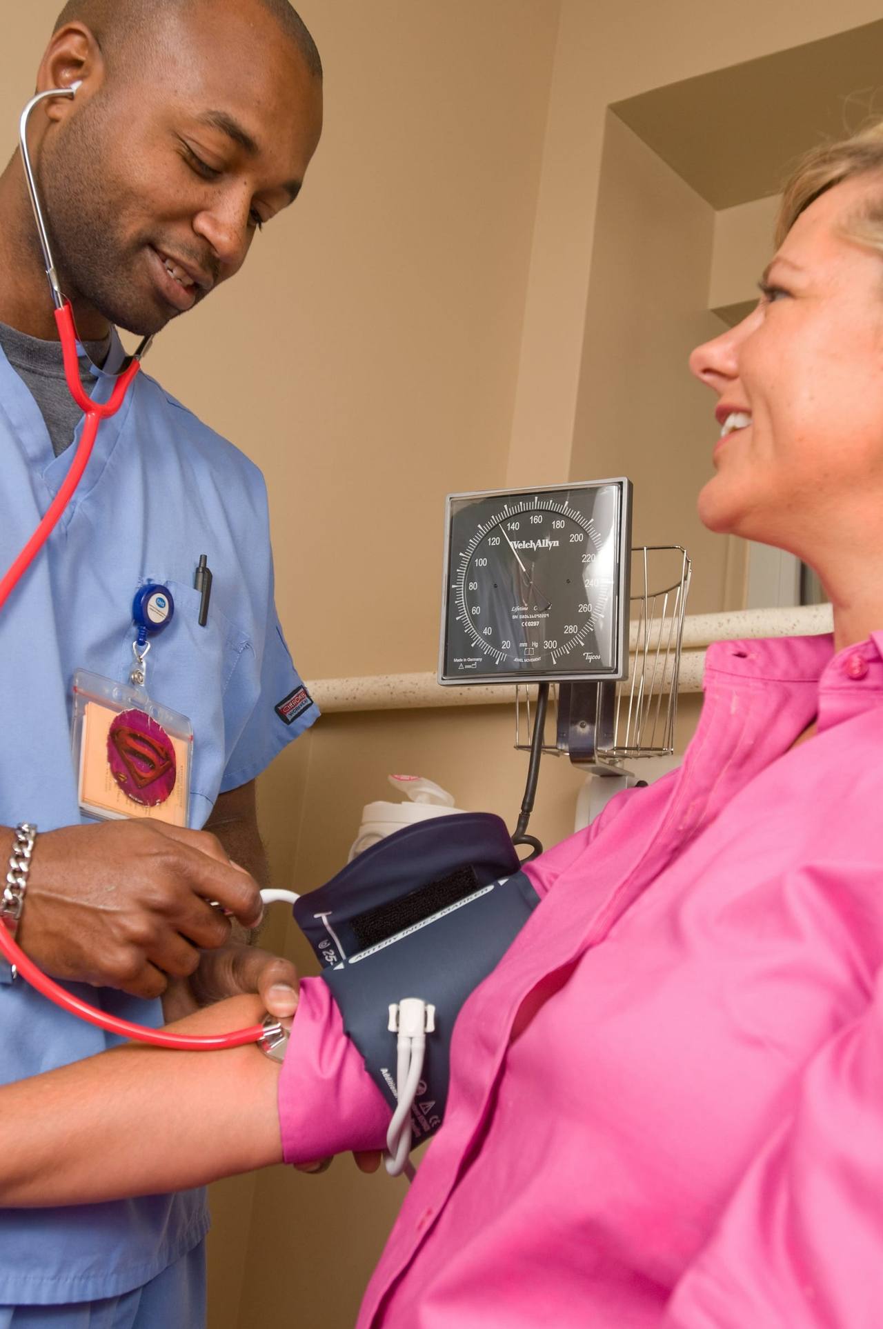 A nurse in blue scrubs takes a smiling woman’s blood pressure in a clinic room
